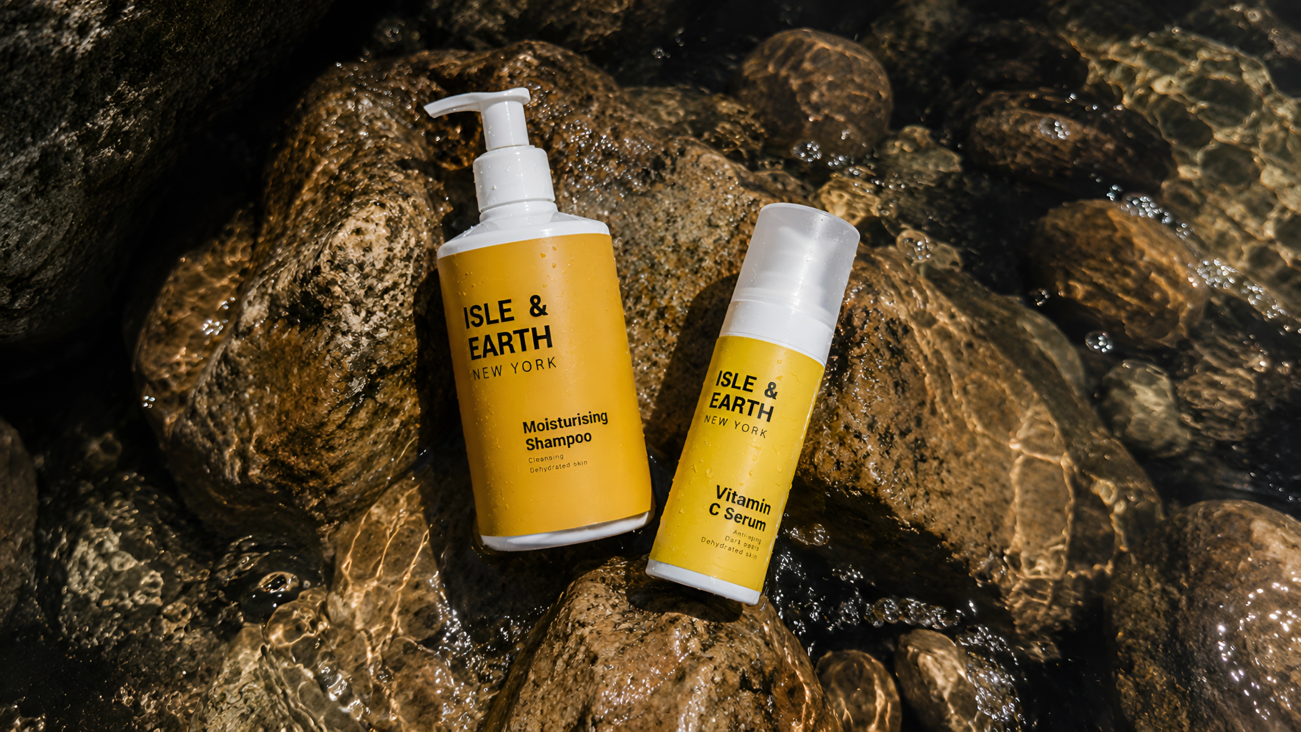 Isle & Earth Moisturising Shampoo and Vitamin C Serum yellow bottles resting on wet coastal rocks with shallow water