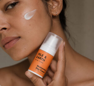Woman applying Isle & Earth Moisturising Day Cream to her cheek with the orange bottle held in hand showing cream on skin