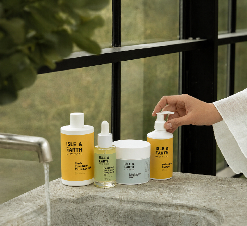 Woman's hand reaching for Isle & Earth Moisturising Shampoo beside Fresh Conditioner Citrus Cocktail, Rosemary Hair Oil, and Icelandic Volcano Scrub on a stone bathroom shelf