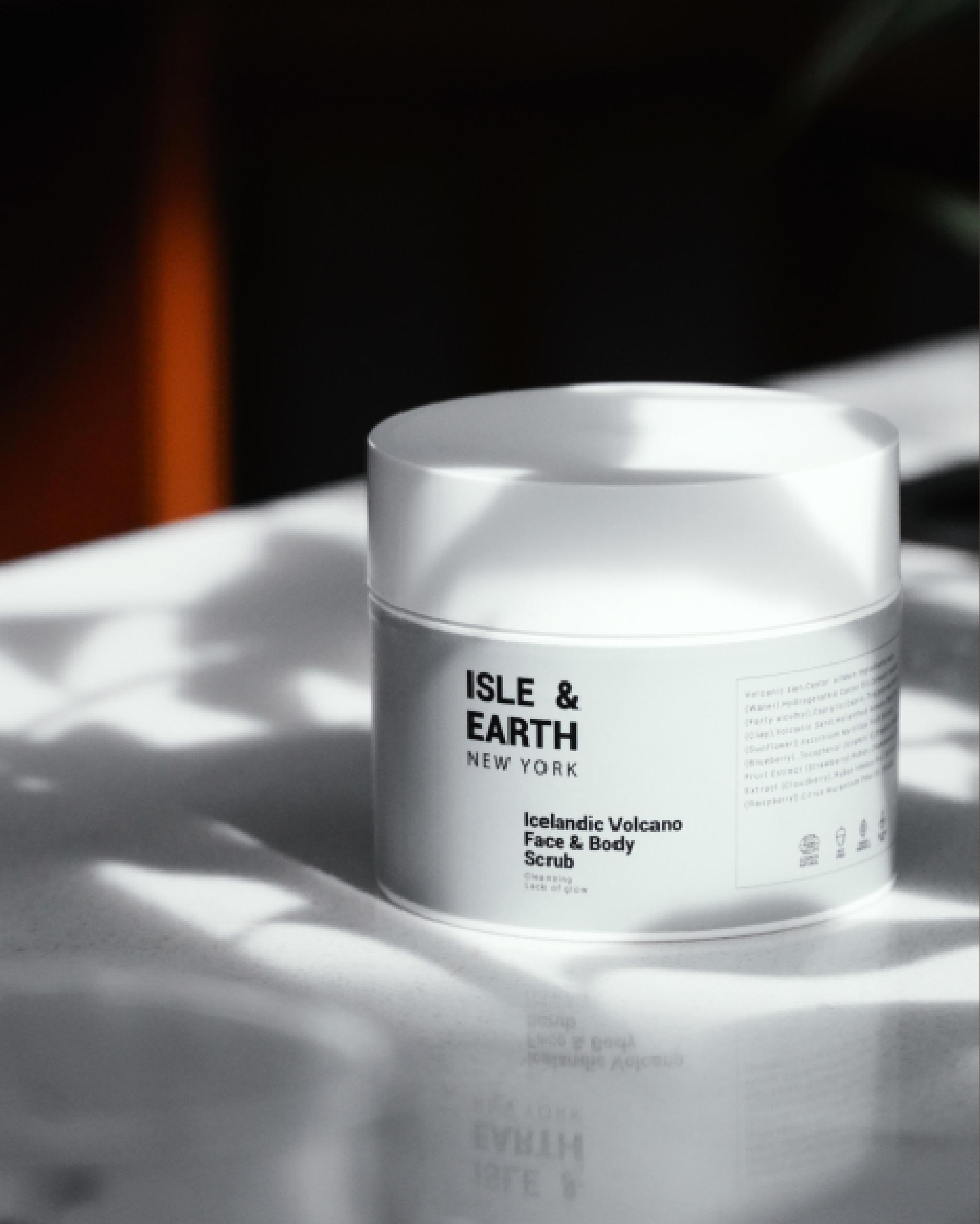 Isle & Earth Icelandic Volcano Face & Body Scrub white jar with sage green label in dramatic dappled natural light on a reflective surface